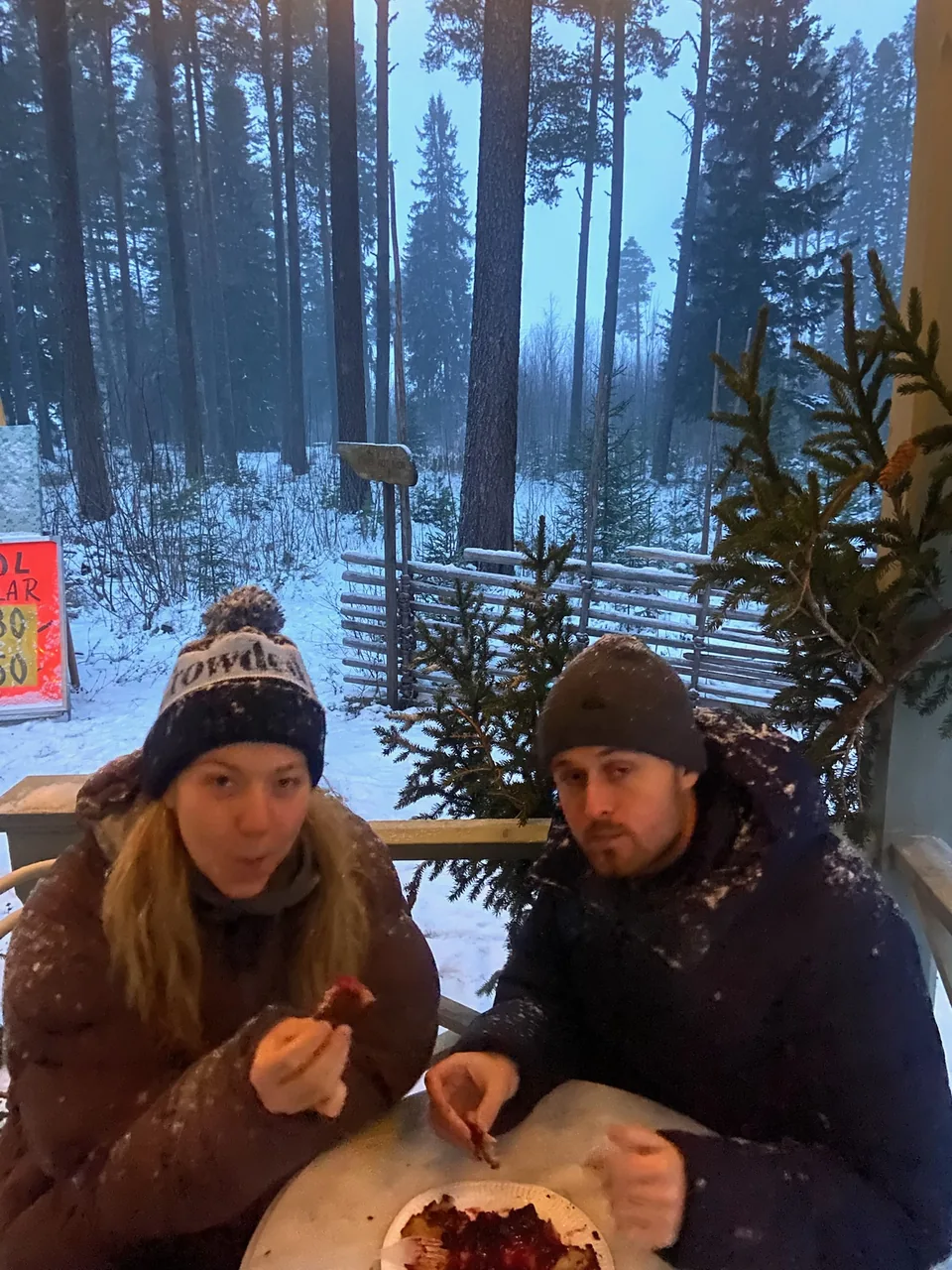 Två i mössor äter våfflor med sylt vid ett bord ute i snöfallet, med granskog bakom.